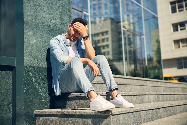 Kind brunette man sitting on the stairs and leaning on the wall while having headache