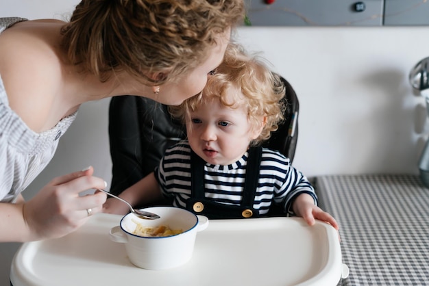 A kind caring mother feeds her young son with useful porridge kisses her forehead