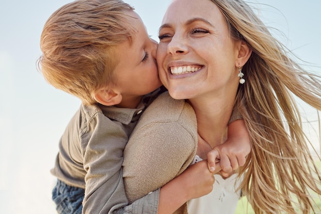 Kiss mother and child with smile in a park for relax adventure and peace together in summer Face of a happy thinking and young mom with her kid kissing and hugging her in nature with a piggyback