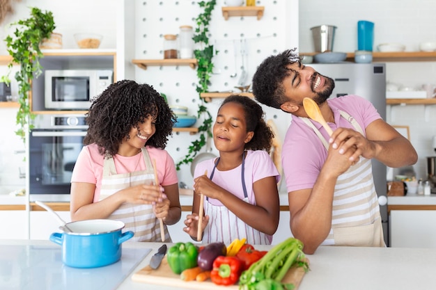 Kitchen Fun Portrait of positive African American parents and little daughter singing and dancing while cooking together at the kitchen using spatula as microphones preparing meal