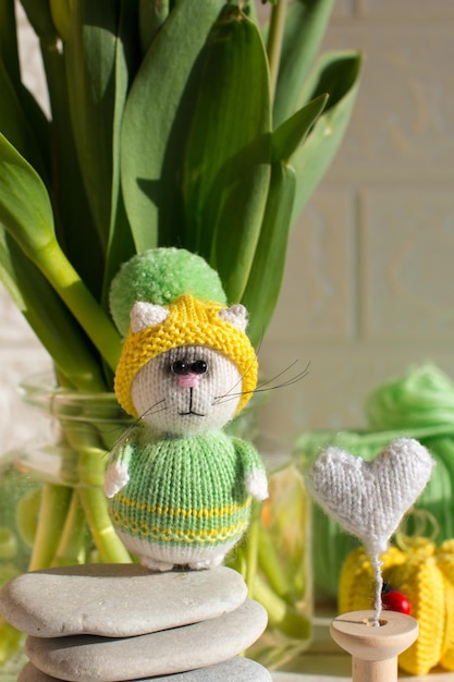 A knitted greenyellow kitten sits with knitting needles under flowers A blooming bouquet of tulips Creative workshop on handmade needlework