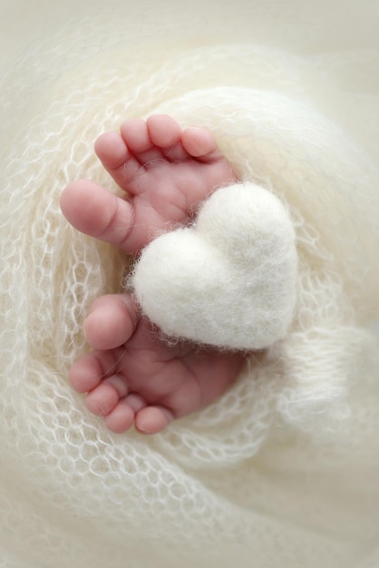 Knitted white heart in the legs of a baby Macro photography The tiny foot of a newborn baby Soft feet of a new born in a white wool blanket Close up of toes heels and feet of a newborn