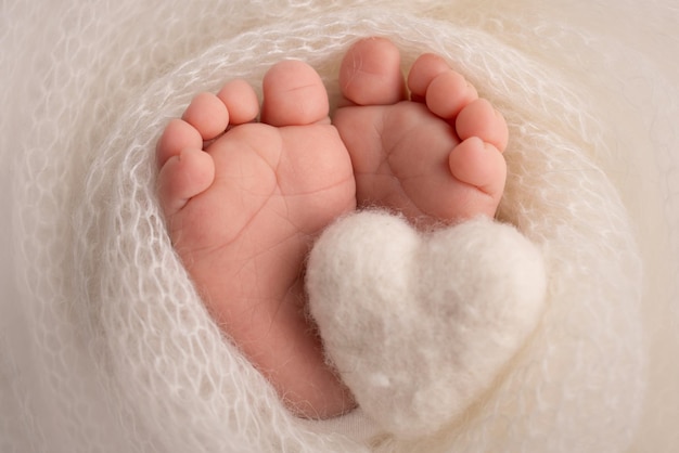 Knitted white heart in the legs of a baby Soft feet of a new born in a white wool blanket Closeup of toes heels and feet of a newborn Macro photography the tiny foot of a newborn baby