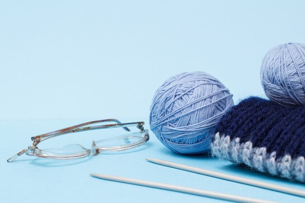 Knitting yarn balls, scarf, metal knitting needles and glasses on a blue background. Knitting concept.