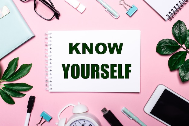 KNOW YOURSELF is written in a white notebook on a pink surface surrounded by business accessories and green leaves.