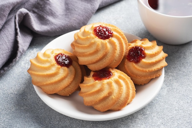 Kurabye shortbread cookies with berry jam in a plate and a cup of tea. Delicious dessert, gray concrete background with copy space.