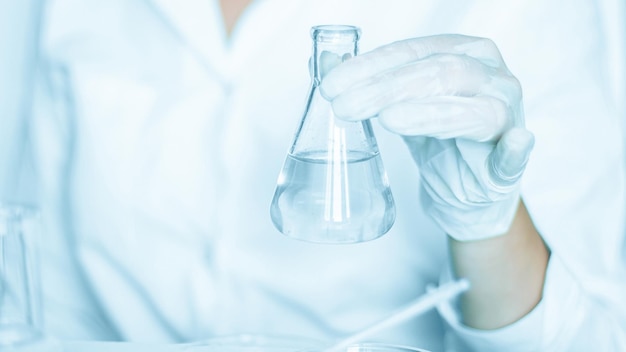 Lab stage Assistant carries out tasks in the laboratory environment Adorned in a lab coat Utilizing glass equipment Located in a blue laboratory Focused on research with transparent liquid