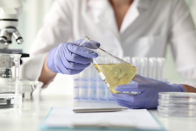 Laboratory technician examines yellow liquid in flask sitting at table with microscope and