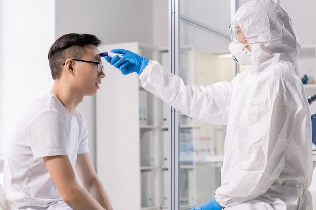 Laboratory worker in protective coveralls, mask, gloves and eyeglasses using special medical tool during examination