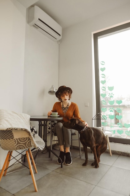 lady in fashionable clothes sits at a table in a cozy cafe holds a cup of coffee in her hand