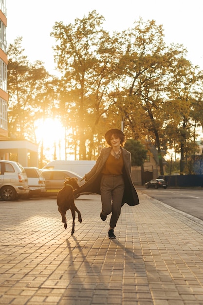 lady in hat and coat running around with playful dog on a leash on the background of autumn trees