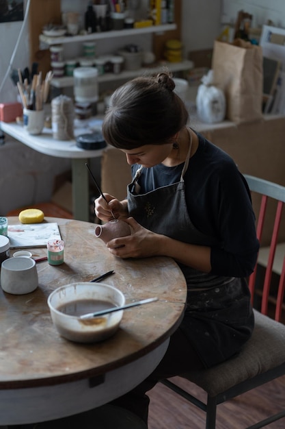 Lady master paints clay jug with serious and concentrated expression sitting at table in workshop