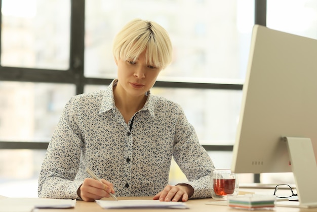 Lady secretary writes notes sitting at wooden table with computer and cup of tea thoughtful