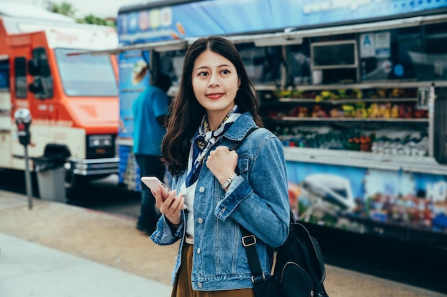 lady studying abroad with smartphone in hand is looking for food truck special recommended online. portrait girl looking away with smile is standing in front of vendor trailers at lunchtime