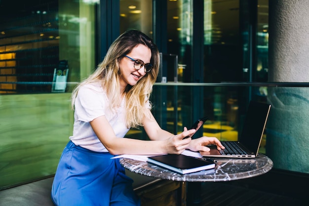 Lady surfing smartphone sitting in cafe with laptop