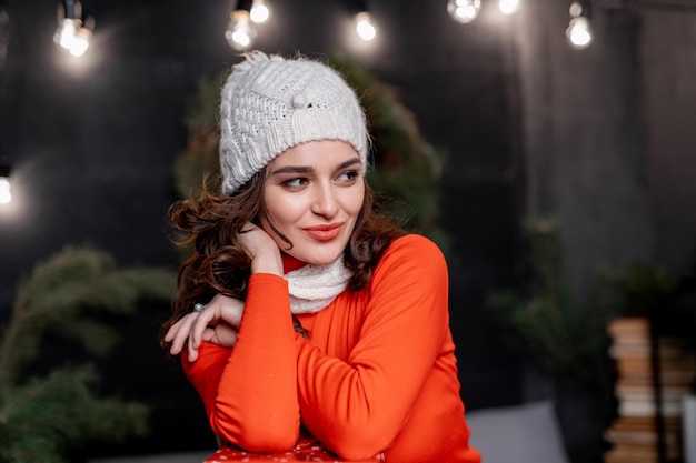 Lady in white knitted hat. Beautiful woman poses to the camera in room decorated before winter holidays. Fest spirit.