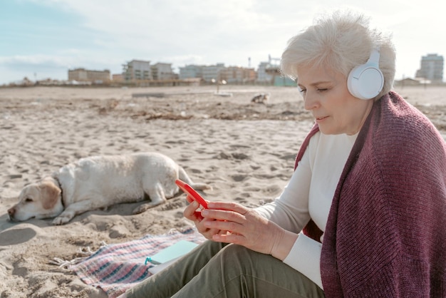 Lady in wireless headphones sitting on the sand beside her dog and staring at the smartphone screen