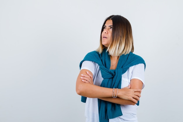 Lady with tied sweater holding arms folded, looking up in white t-shirt and looking pensive , front view.