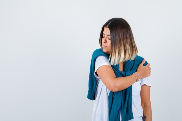 Lady with tied sweater keeping hand on arm, looking down in white t-shirt and looking desperate. front view.