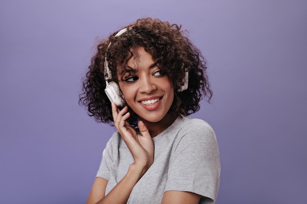 Lady with wavy hair listens to music in white headphones Attractive woman in gray tee laughing on purple isolated backdrop