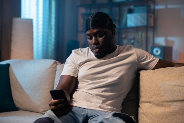 A laidback darkskinned man in a white tshirt sits on a sofa at night in an apartment