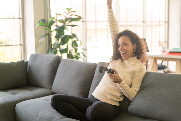 Laitin woman playing videogames with hands holding joystick on sofa at home