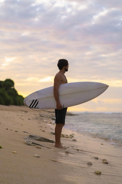 Landscape image of male surfer busy walking on the beach at sunrise while carrying his surfboard under his arm with the ocean waves breaking in the background. Young handsome male surfer on the ocean