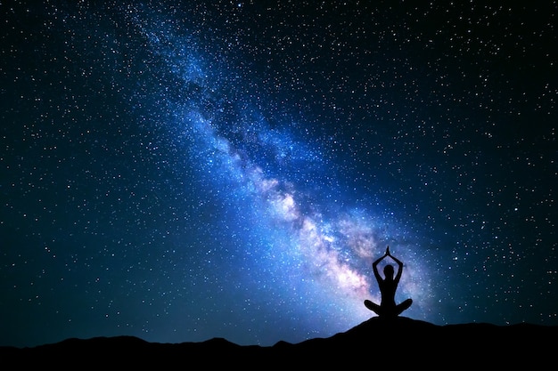 Landscape with blue Milky Way. Night sky with stars and silhouette of a girl practicing yoga in the hill.