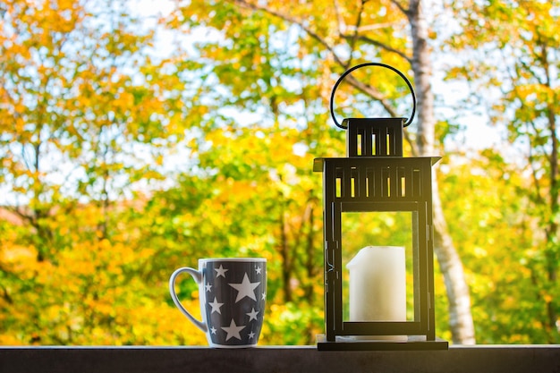 The lantern with a candle and a cup of coffee on a background of autumn forest