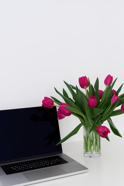 Laptop and bouquet of fresh tulip flowers in a vase on a white table