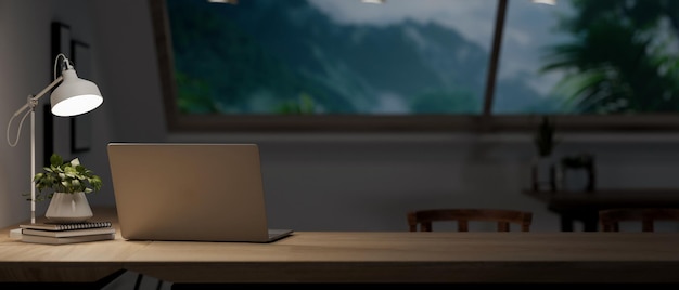 A laptop computer books a decor plant and a table lamp on a wooden table in a dark room