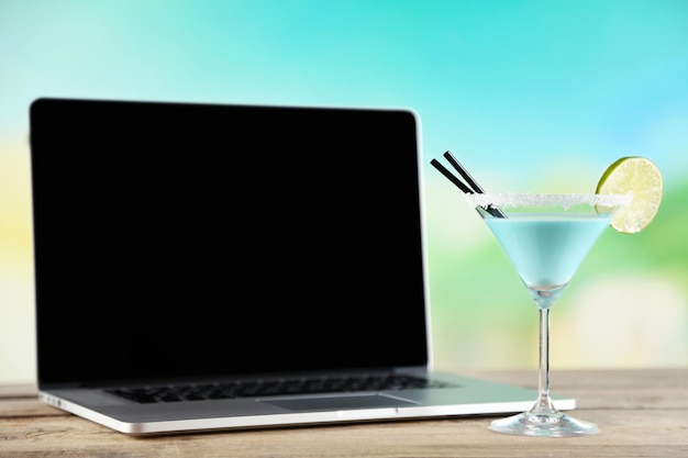 Laptop and glass of summer cocktail on wooden table on bright blurred background