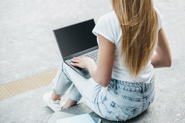 Laptop mockup. Young student girl uses laptop with white screen, sitting on stairs and looking at empty black monitor screen near office building. Copy space for design