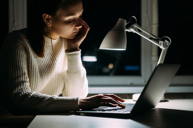 Laptop searching Boring woman Internet connection Distance work Tired lady leaning hand scrolling notebook in table lamp light dark night room interior