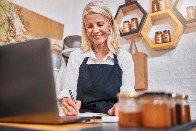 Laptop small business and senior woman writing notes while doing inventory or online marketing Happy smile and elderly entrepreneur in a honey store working on admin or shop sales with a computer