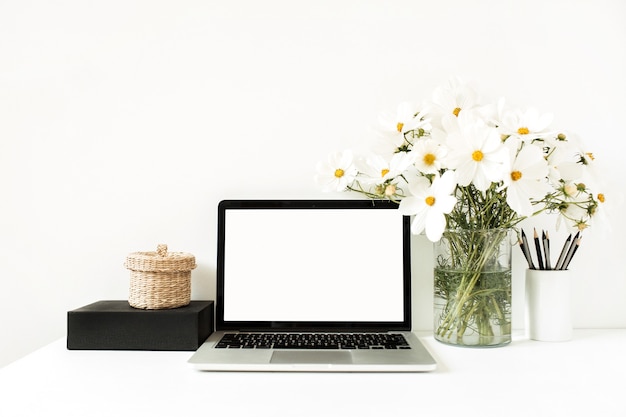 Laptop standing on white table against white wall with daisies in vase, black box, straw basket.