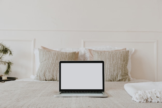 Laptop with blank copy space screen display in bed with plaid, pillows against white wall. Aesthetic morning composition.