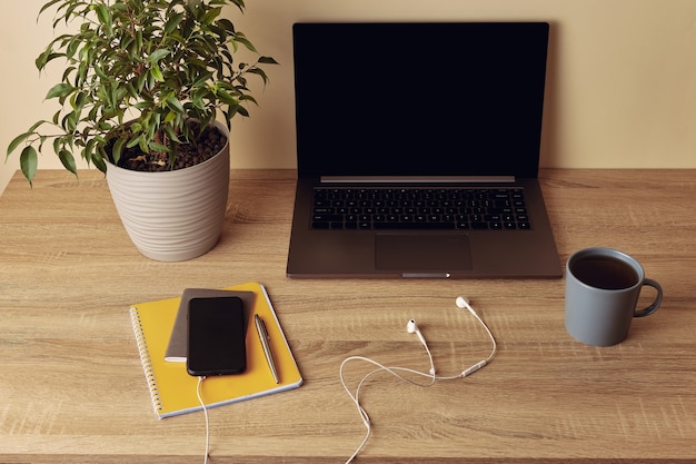 Laptop with blank screen, potted plant, mug, yellow notepad, pen, mobile phone and headphones.