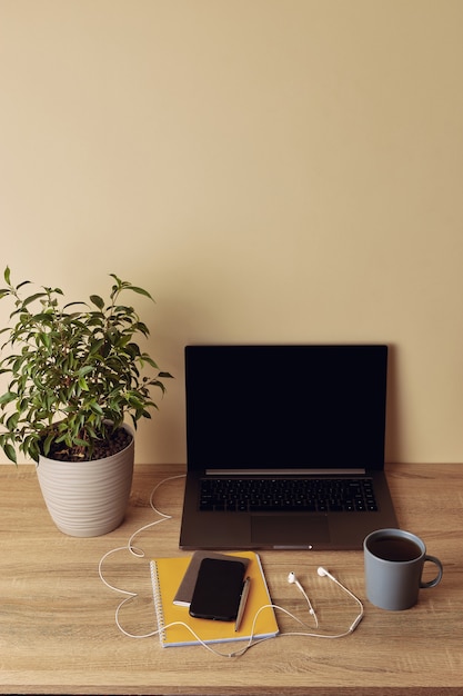 Laptop with blank screen, potted plant, mug, yellow notepad, pen, mobile phone and headphones.