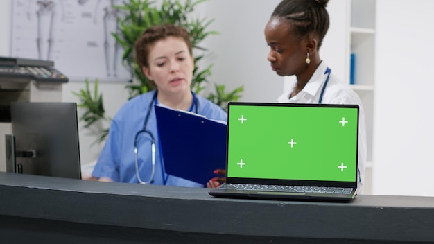 Laptop with greenscreen display on hospital reception counter used by medical team. Working with blank copyspace template, isolated mockup and chroma key background on pc computer.