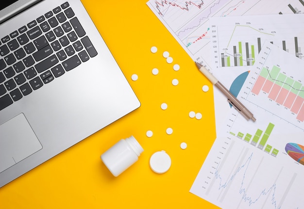 Laptop with pills bottle,  graphs and charts on a yellow background. Business plan, financial analytics, medicial statistics.