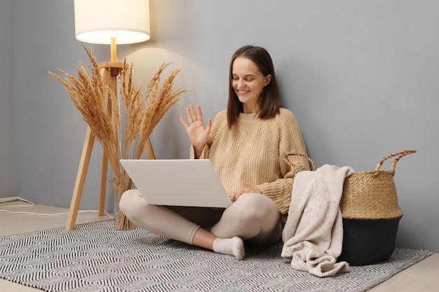 Laptop work internet Optimistic friendly brown haired woman wearing beige sweater working on computer while sitting on floor in home interior waving hand greeting has video call