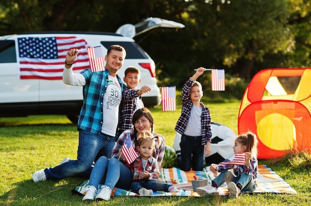 Large american family spending time together. With USA flags against big suv car outdoor. America holiday. Four kids.