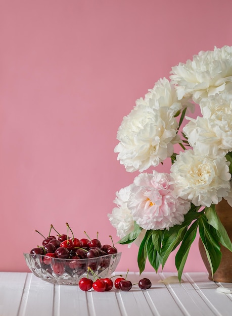 A large bouquet of peonies in a ceramic vase on the table cherries in a bowl