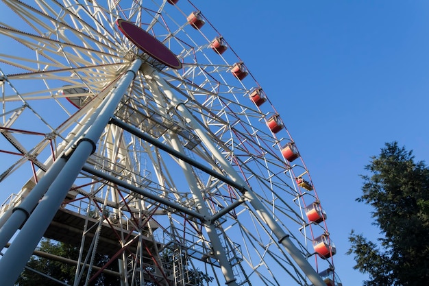 A large ferris wheel in an amusement park with red and yellow cabins on a blue sky background trees