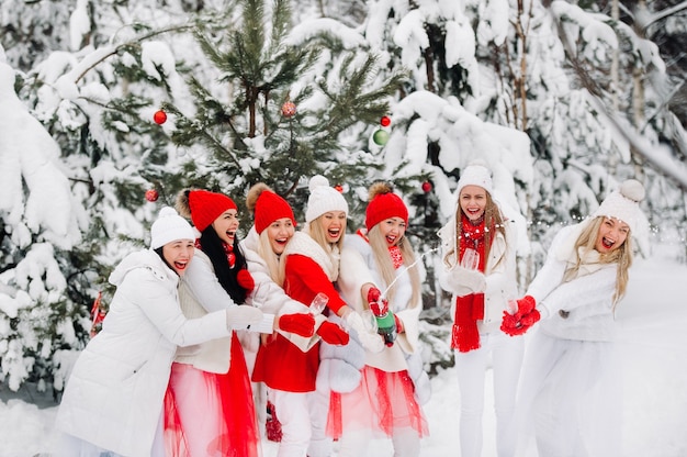 A large group of girls with glasses of champagne in their hands stands in the winter forest.Girls in red and white clothes with new year's drinks in a snow-covered forest.