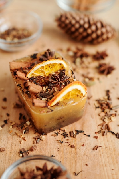 Large handmade soap bar with cinnamon sticks, star anise and orange slices on wooden table with grated spices near by