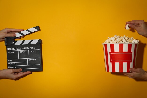 Large paper cup of popcorn and clapperboard in hands on yellow background space for text