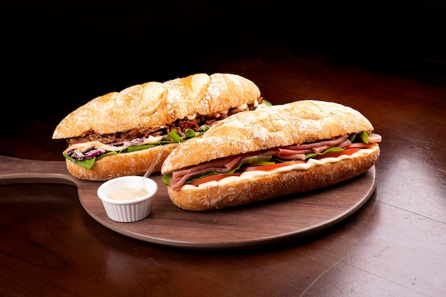 Large smoked carpaccio sandwich with arugula tomato and gruiere cheese on italian baguette and brisket beef on wooden round board and dark background with sauces from above