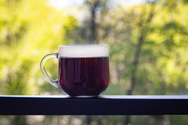 A large transparent cup with black coffee stands on a dark iron table against a blurred background of summer green trees. Good morning concept. Fresh ideas. Soft focus.